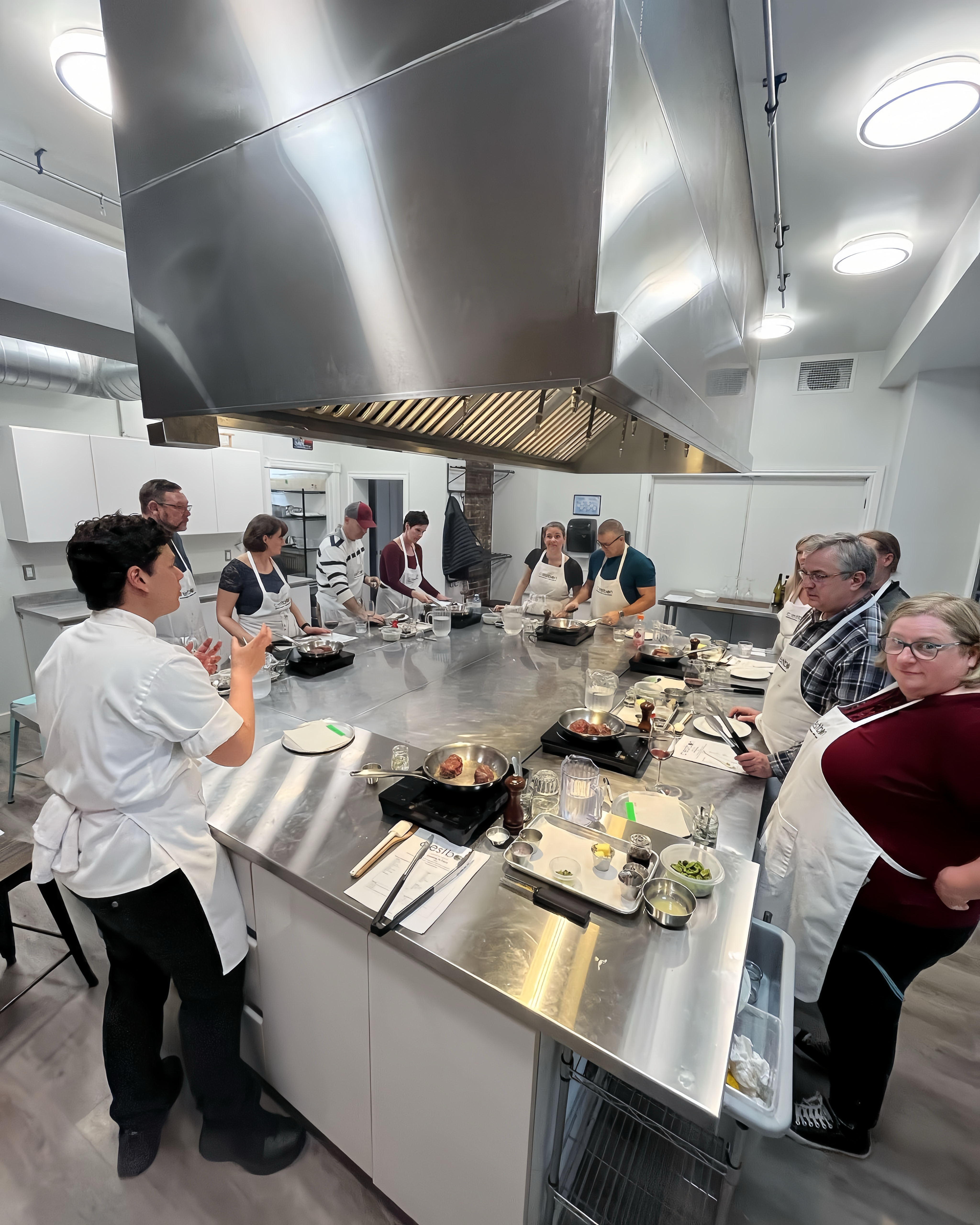 A professional chef instructor in front of 5 couples standing around a central hub in an professional kitchen, learning how to cook.