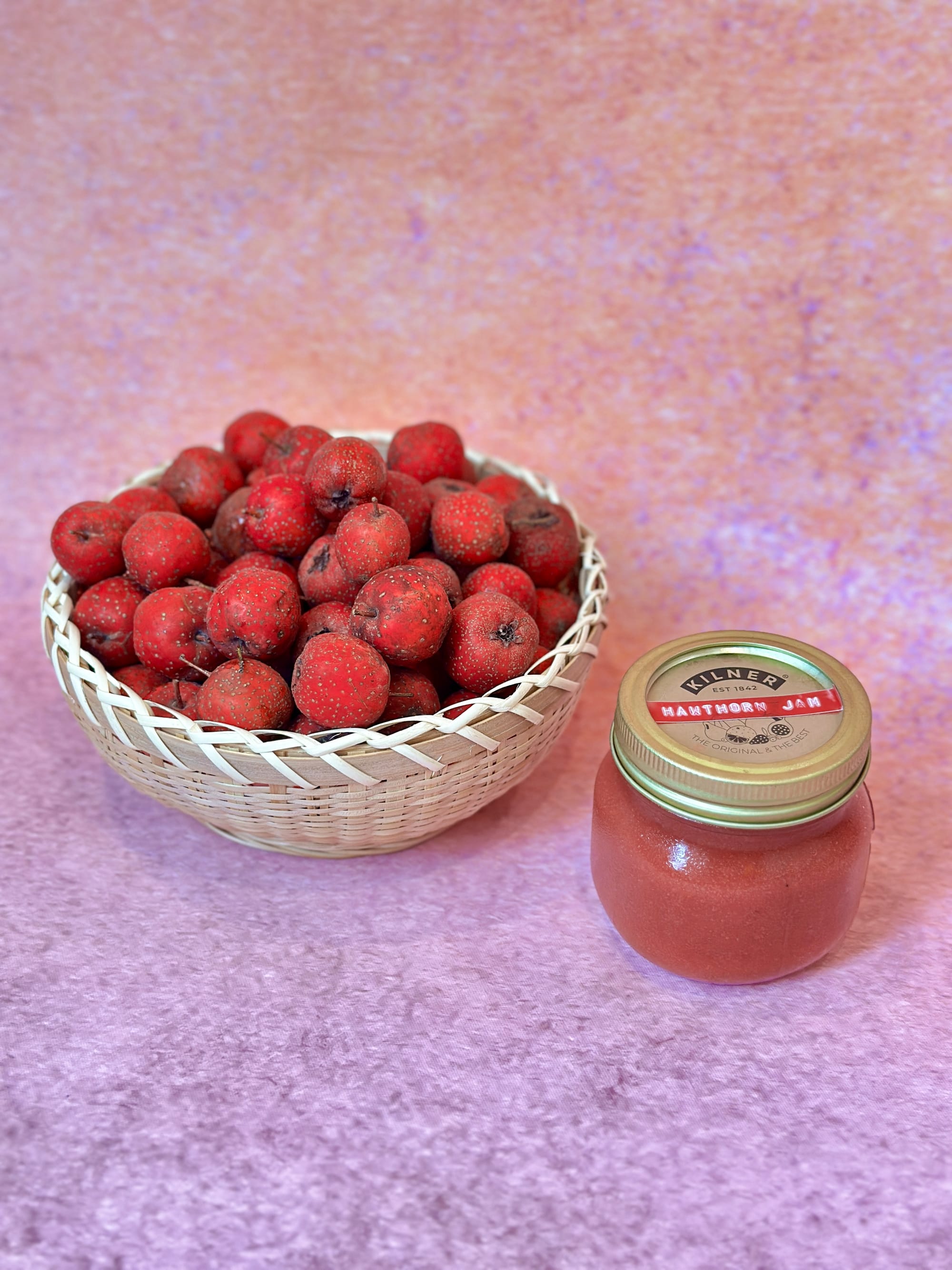 Red hawthorn berries in a bamboo basket next to a small jar of hawthorn jam.