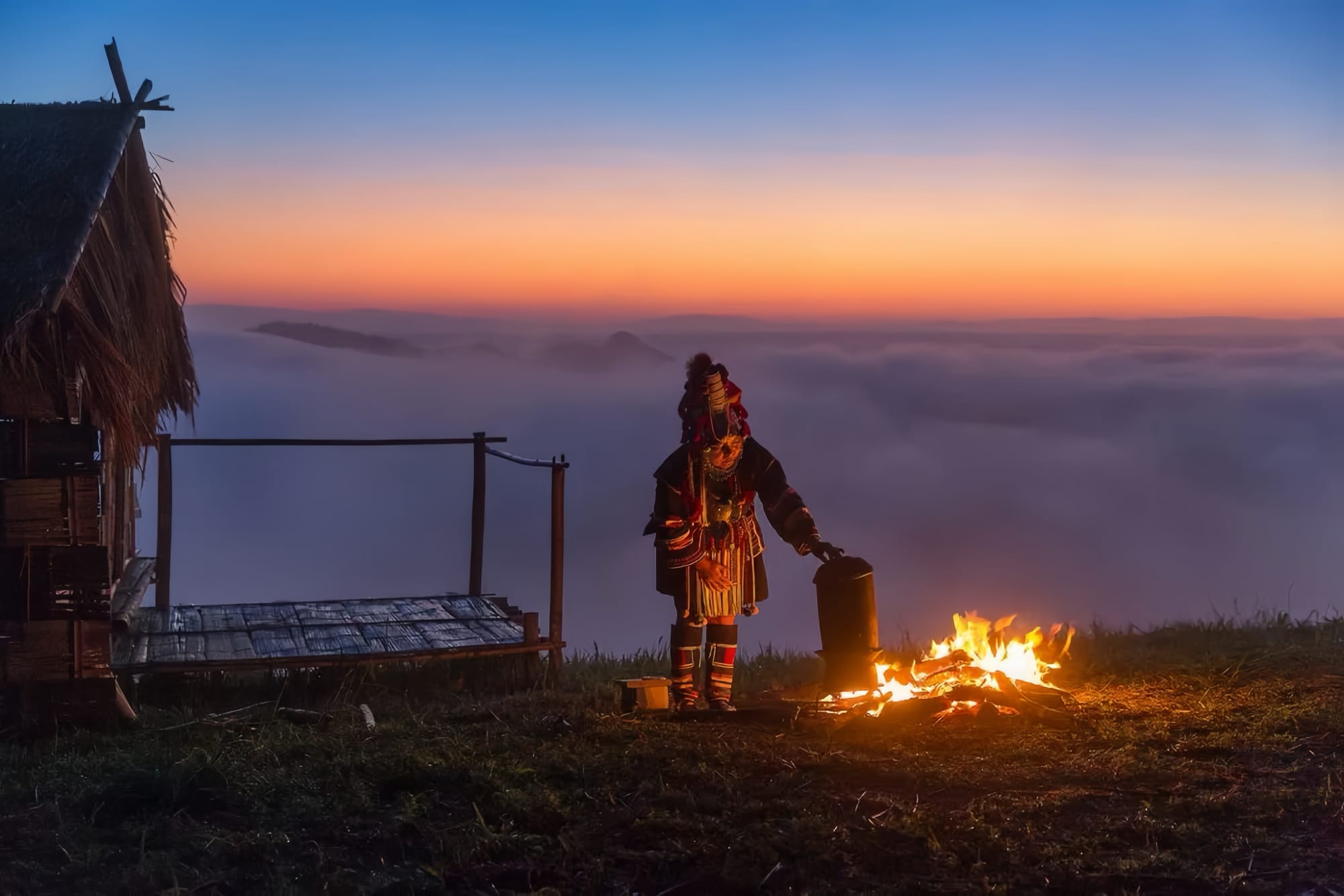 A traditionally dressed Hani woman at the top of a grassy mountain, tending to a fire in the evening next to the wooden porch of a traditional cabin.