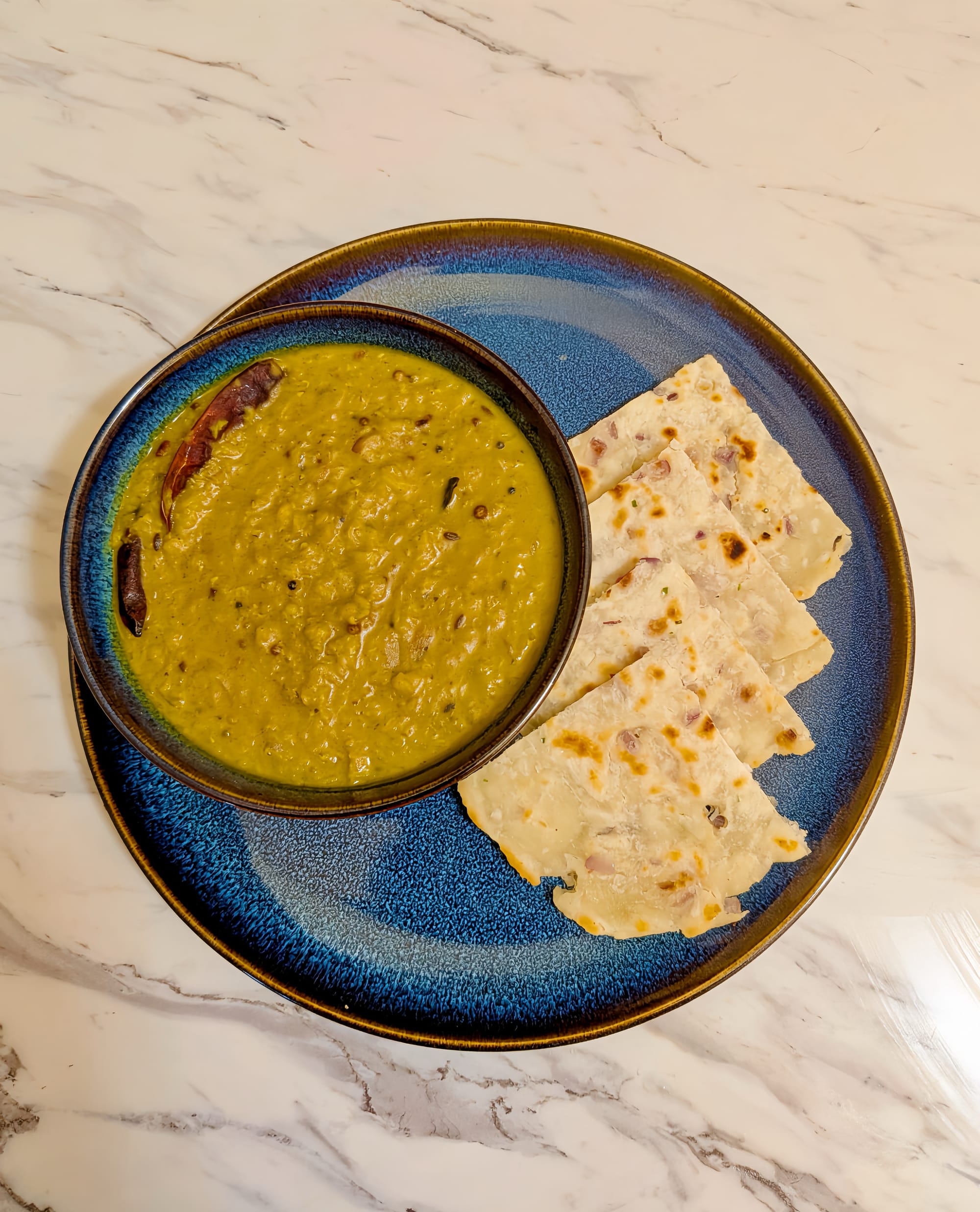 On a marble background, a blue plate with triangular cut pieces of Pol Roti next to a bowl of yellow/green Dhal Curry