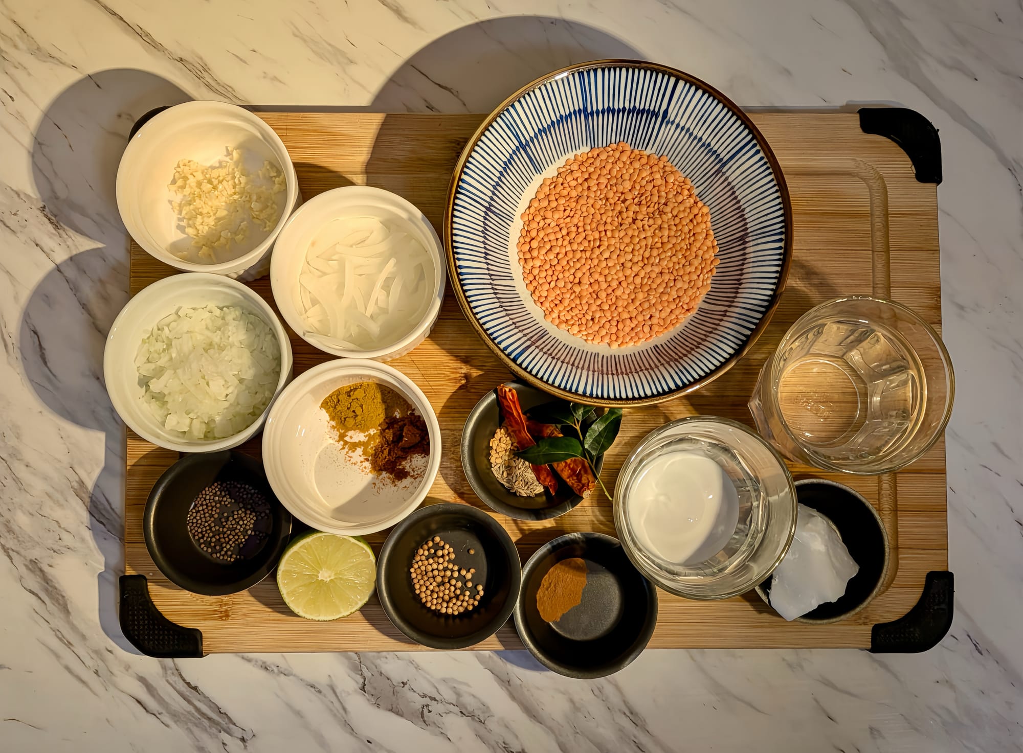 On a marble backdrop, a bamboo cutting board holds small dishes with the ingredients measured out for Dhal Curry