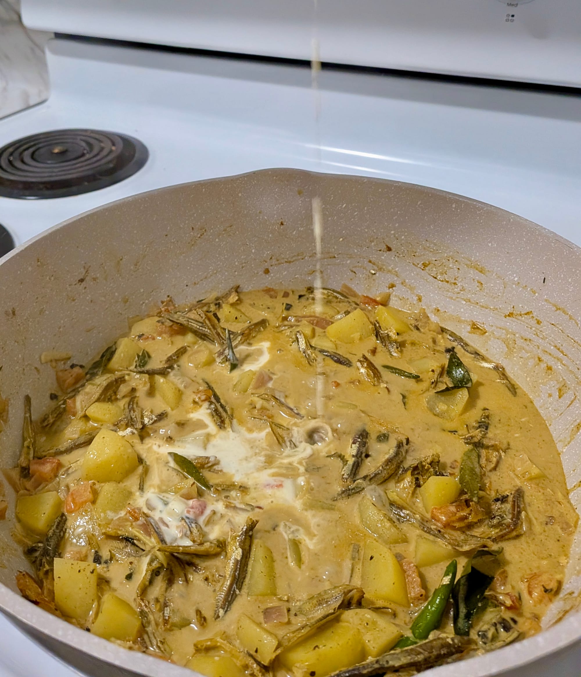 A golden yellow breakfast curry with potatoes, chilis, and other ingredients in a pot on a white stove.