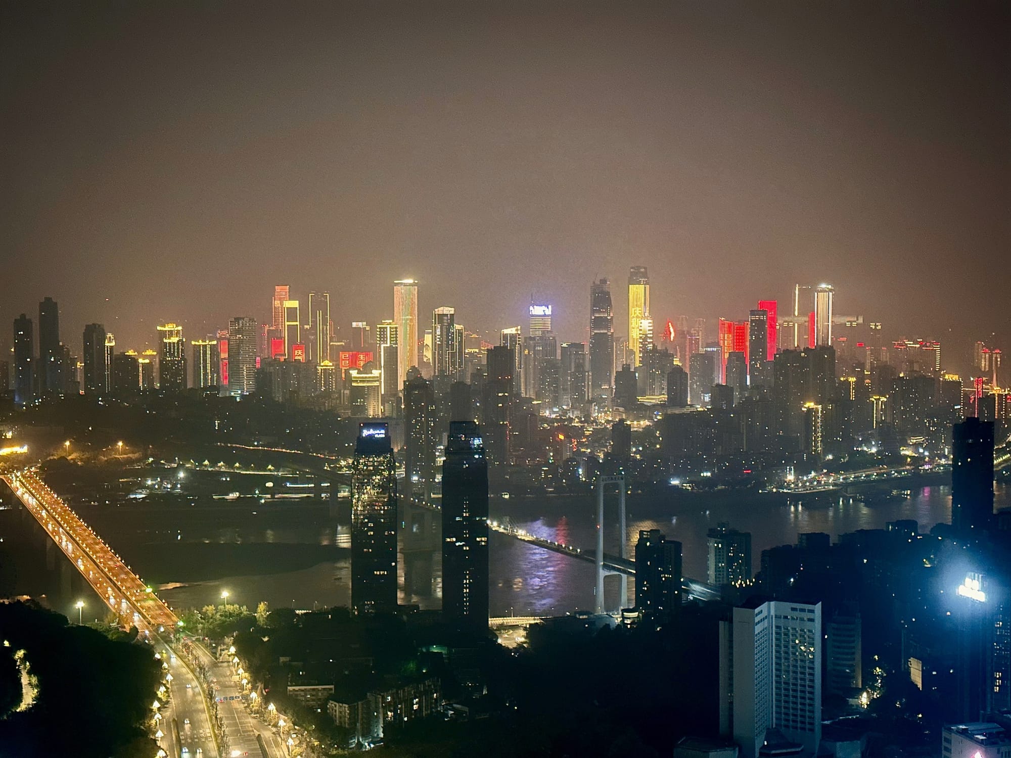 Chongqing cityscape at night, featuring two bridges across a river cutting the city in half. Across the river, brightly lit golden and red skyscrapers make the sky glow a faint gold color.