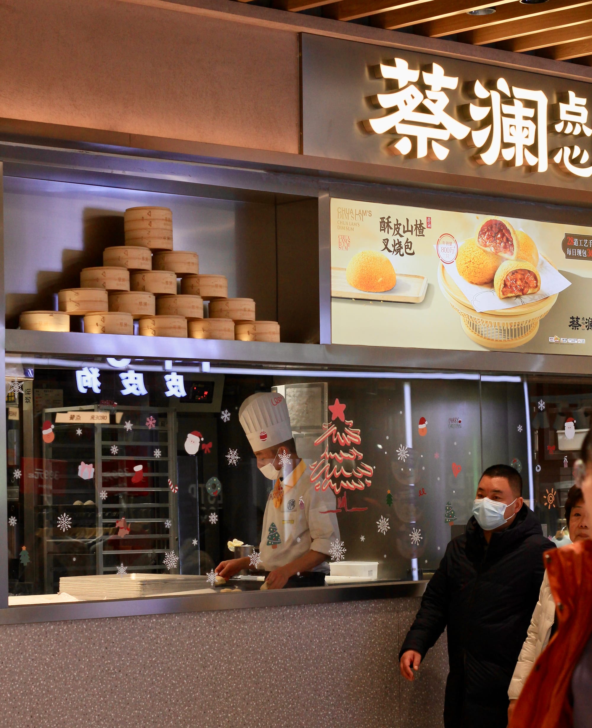 A small bakery has a glass viewing window decorated with Christmas drawings. A chef behind the glass prepares pastries as customers walk past.