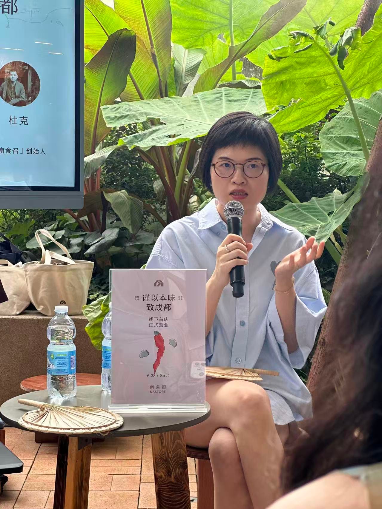 Tianluo, a famous food writer in China, sits at a small coffee table featuring an advertisement for her book. She is holding a microphone and speaking to a small audience at a book signing event in Chengdu.