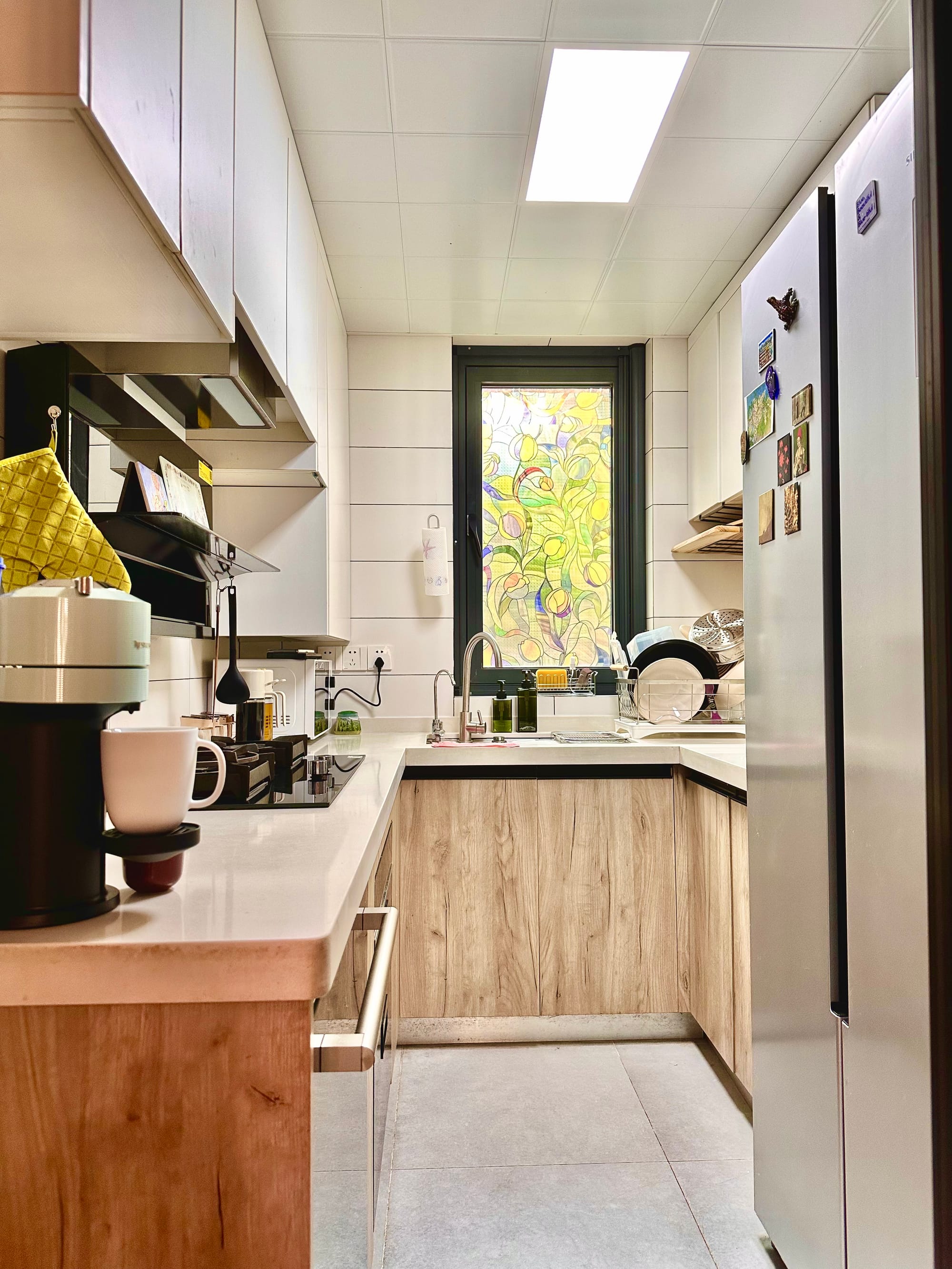 A narrow kitchen space with a counter and stove-top on the left and a Nespresso machine. On the right, a silver fridge. Straight ahead, a sink with a stained glass window above.