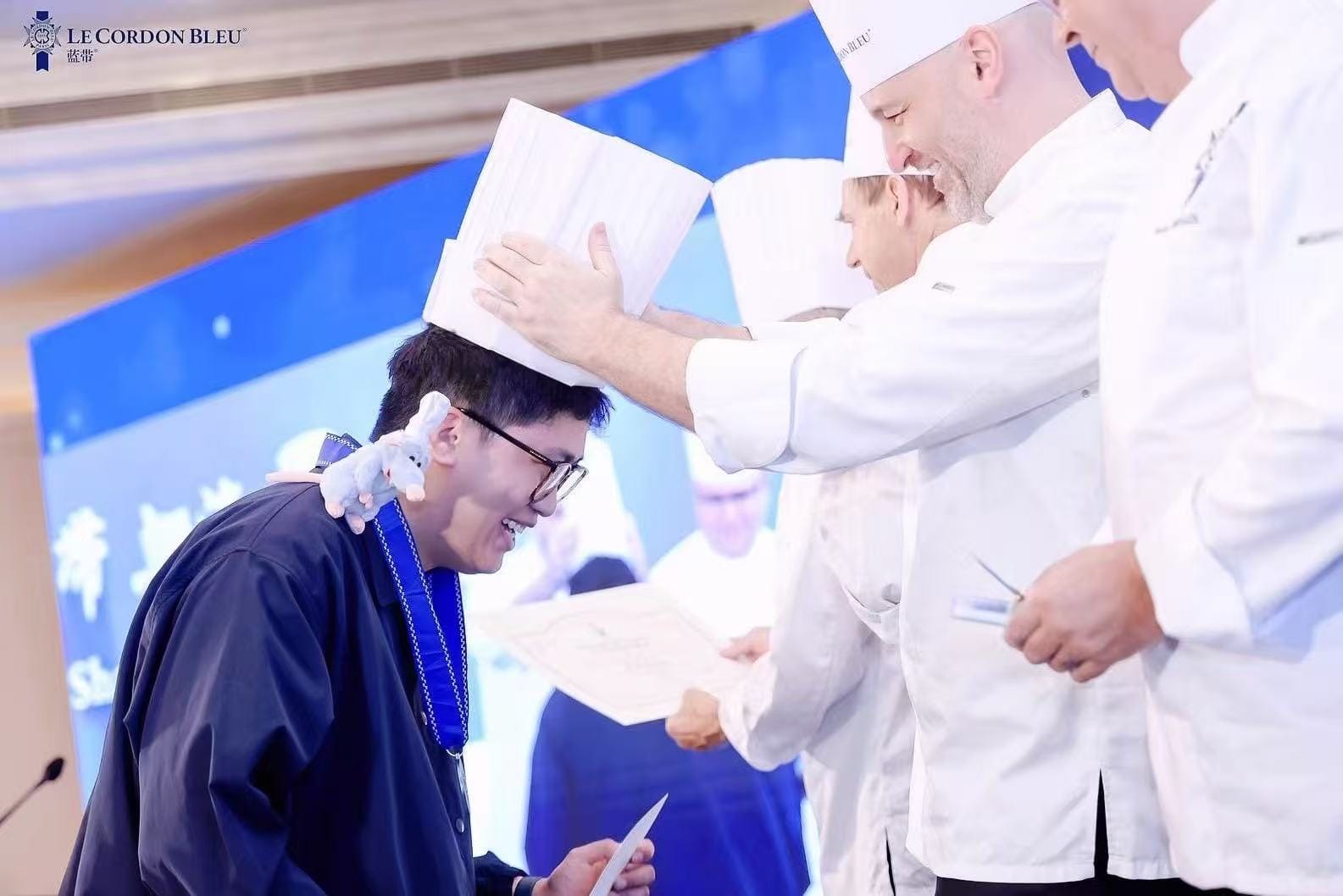Wei Yuan receives his chefs hat during his graduation from Le Cordon Bleu, Shanghai with a stuffed toy of Remy on his shoulder.
