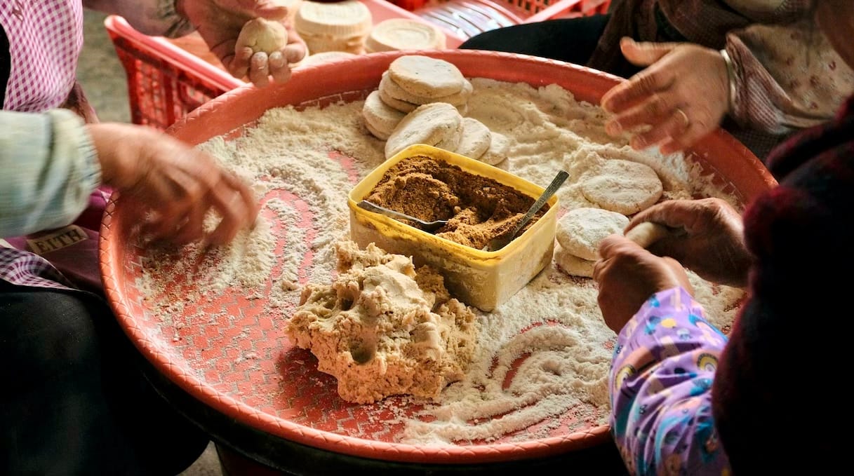 Three elderly women sit around a plastic tray hand-rolling brown sugar into sweet cakes.