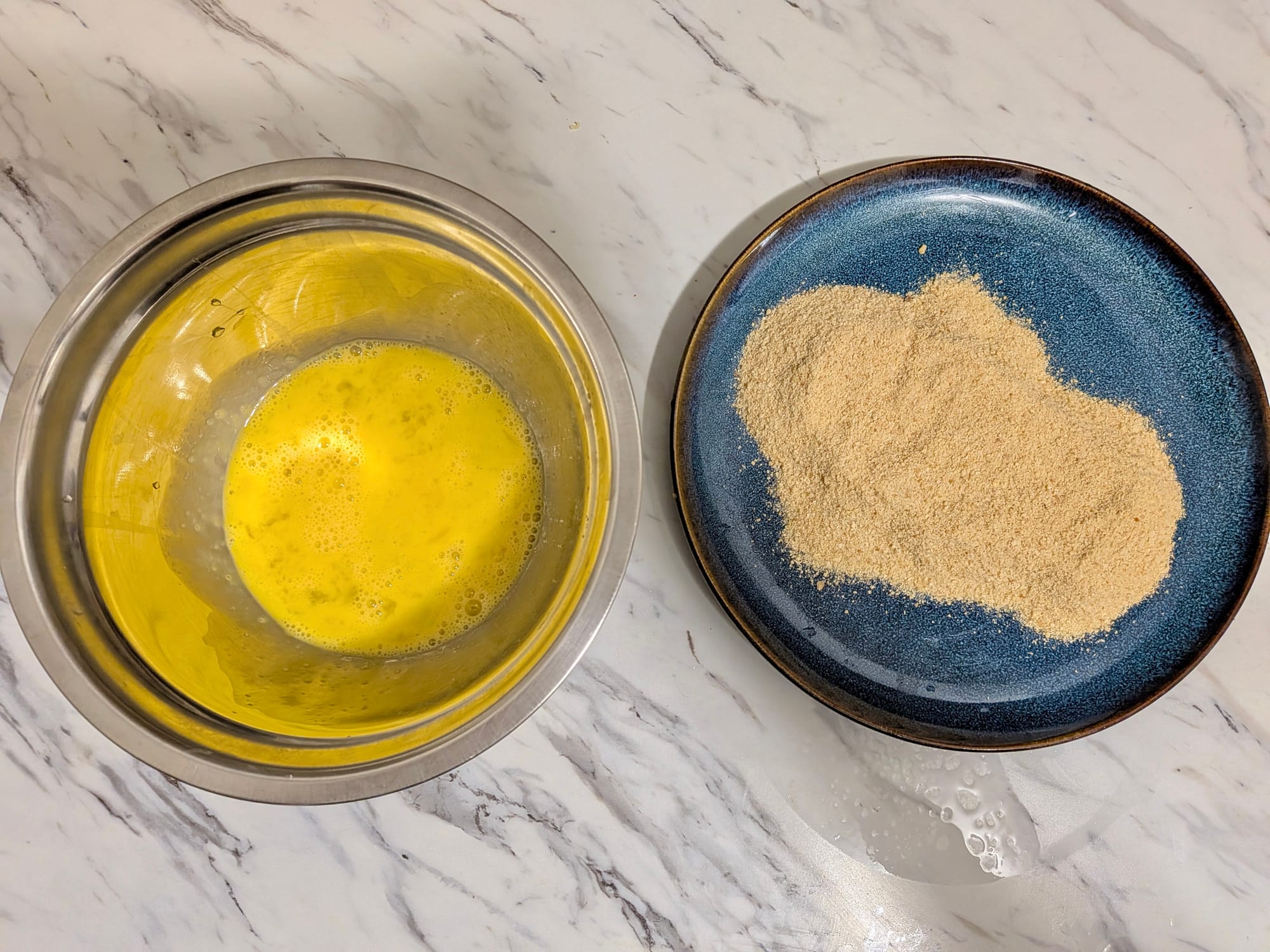 A stainless steal bowl with egg-wash and a blue porcelain plate with breadcrumbs for frying chicken rolls.