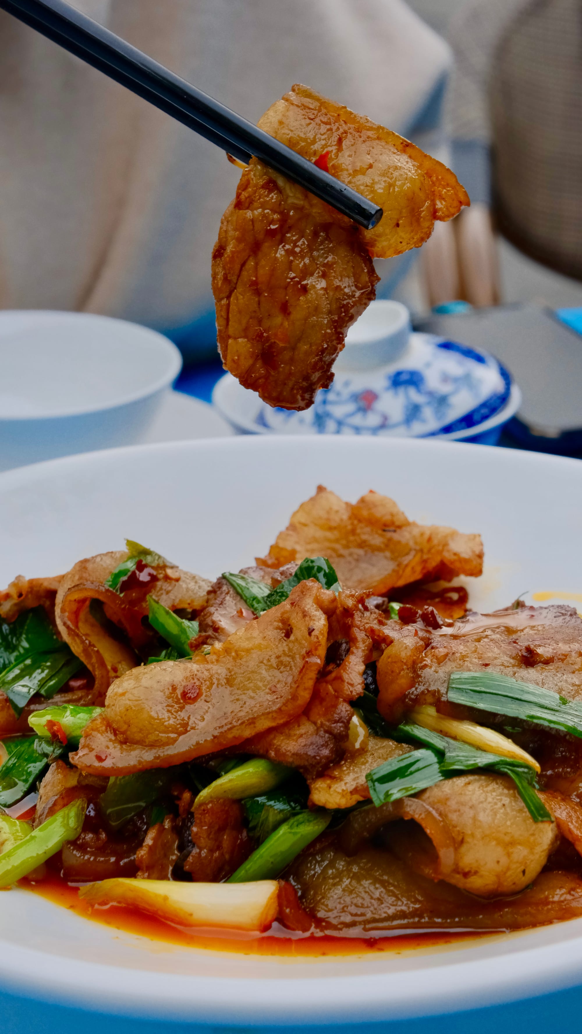 回锅肉 huí guō ròu, Twice-Cooked Pork, sits on a white dish. Thinly sliced pork with a redish-orange sauce and green onion chutes.
