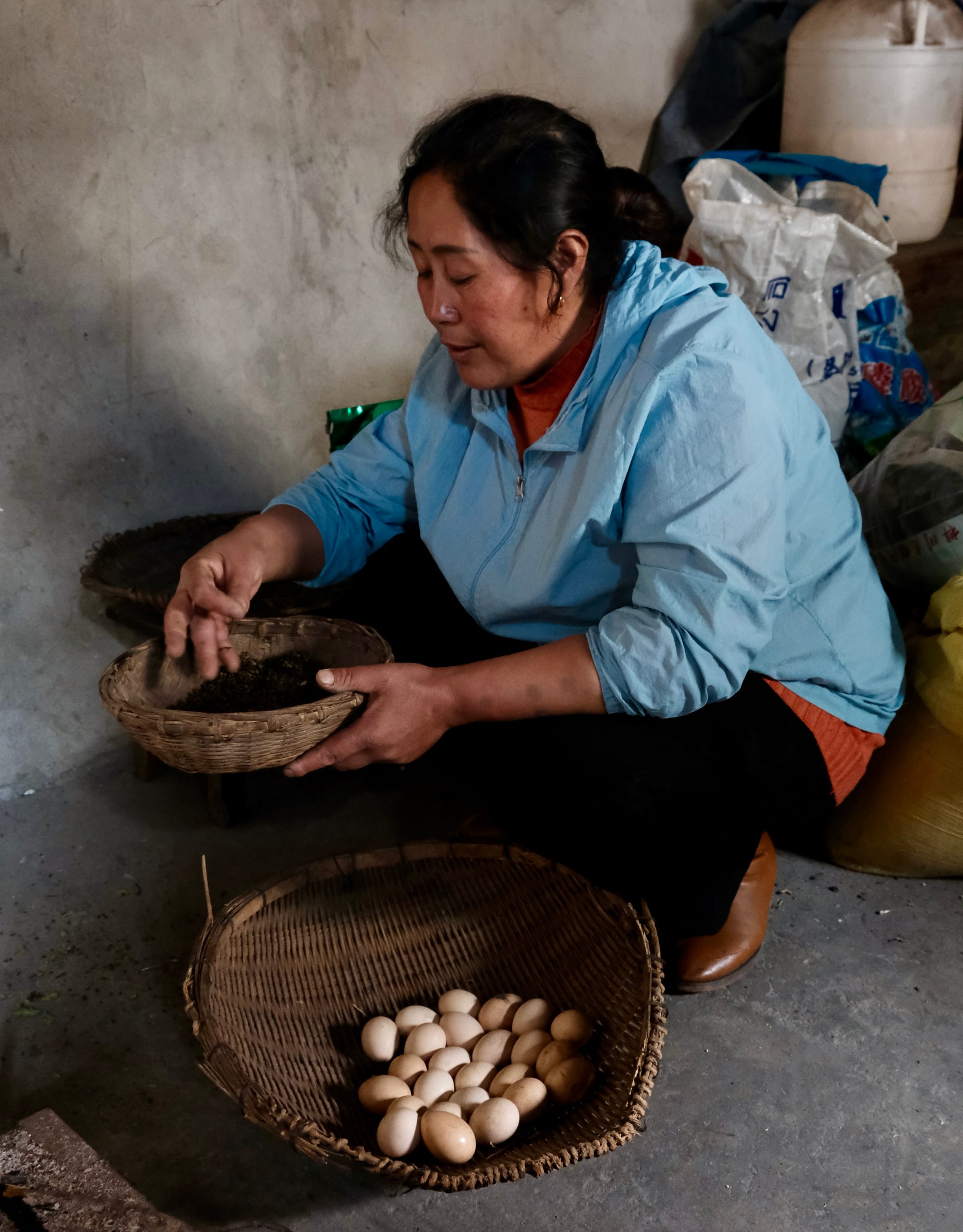 An elder Yi lady dressed in a blue top crouches down sorting pickled greens next to other produce.