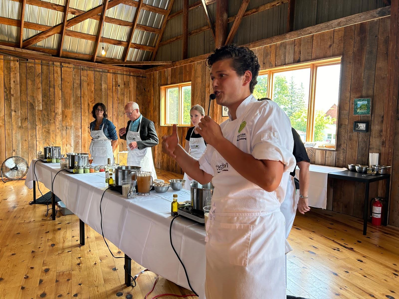 Arnold standing in front of a demonstration table setup with hot plates and pots and sets of ingredients, leading a cooking class as four volunteers stand behind waiting for instructions.