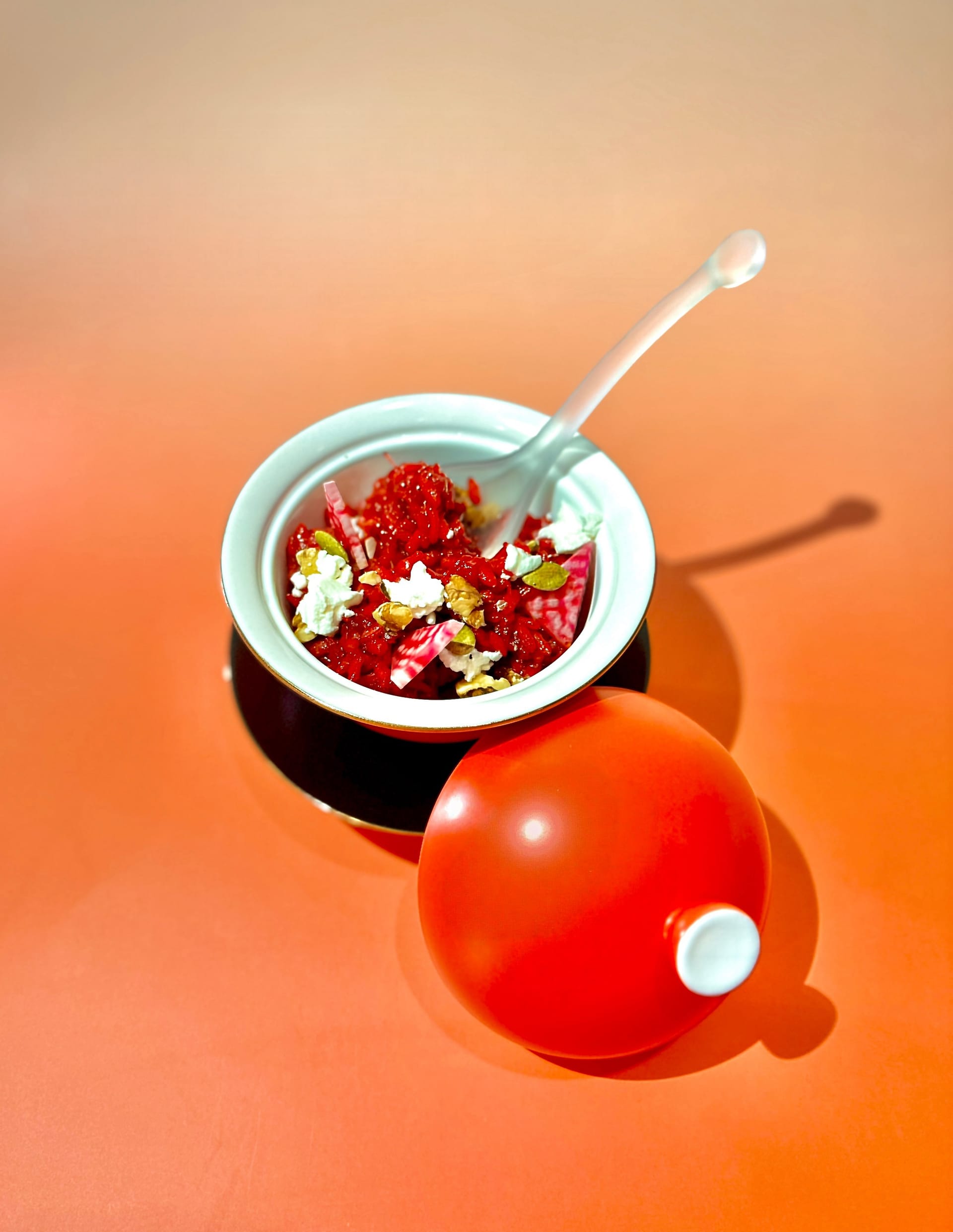 A black porcelain dish, with a red and white bowl filled with red beet risotto, with a clear glass spoon and a red porcelain lid propped up against the bowl.