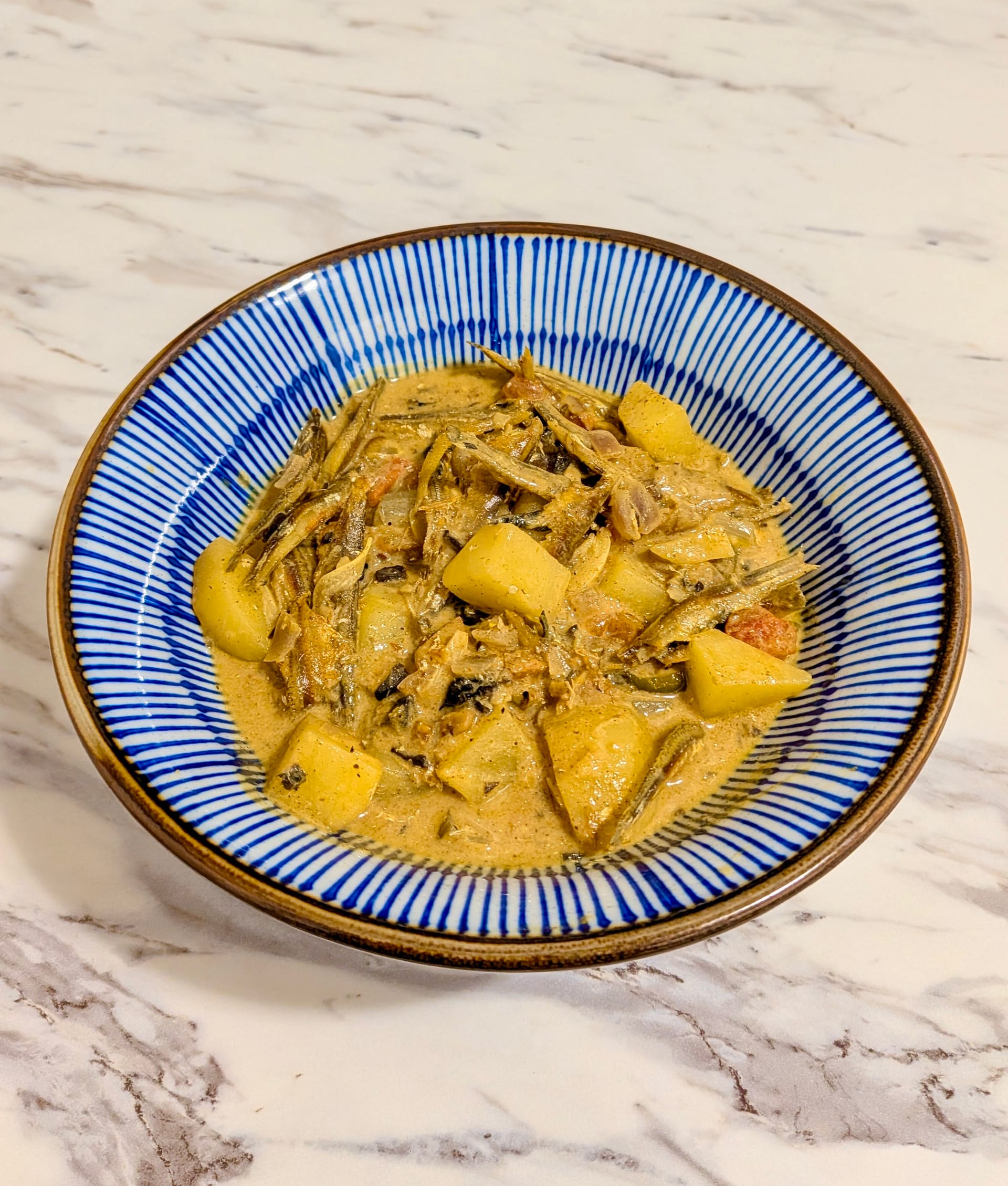 A decorated bowl on a white countertop with a golden yellow potato and sprat curry.