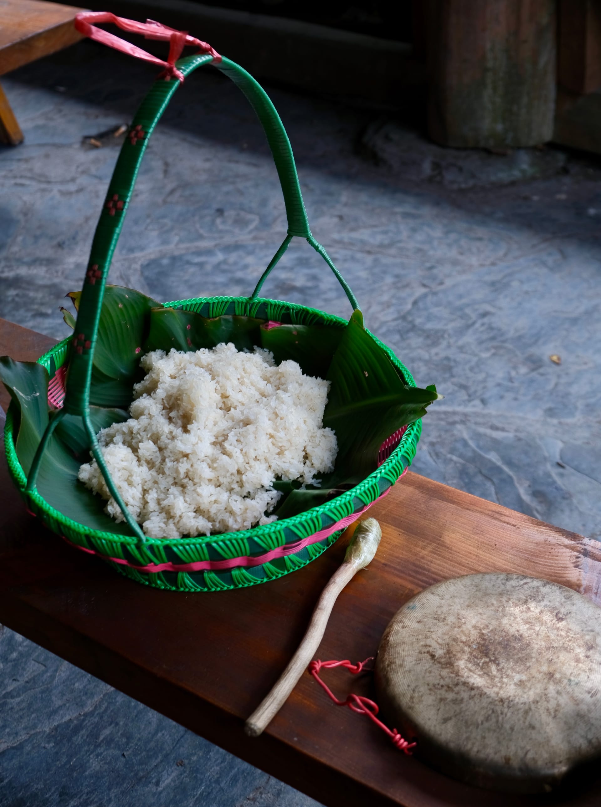 A colorful green and pink basket filled with glutinous white rice sits on a wooden bench.