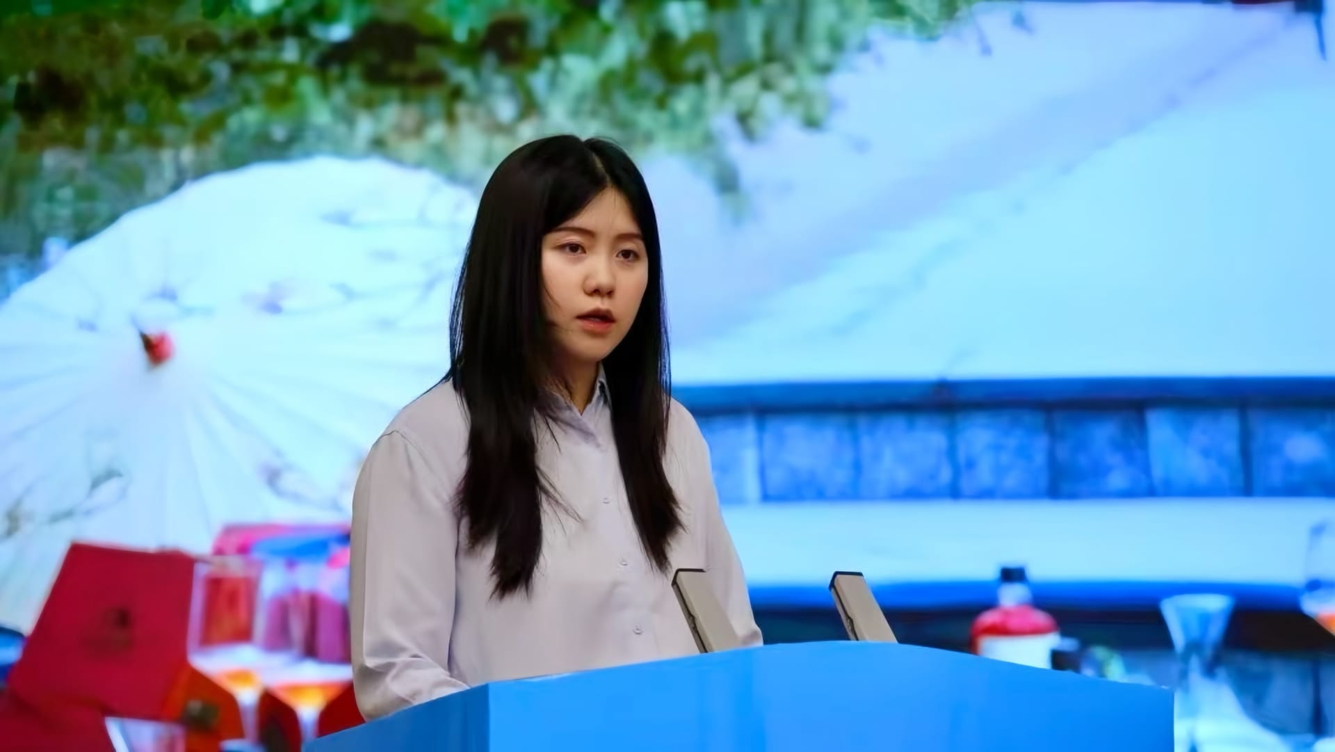 Yifei Guo speaking at a sky blue podium, with a Chinese market behind her.