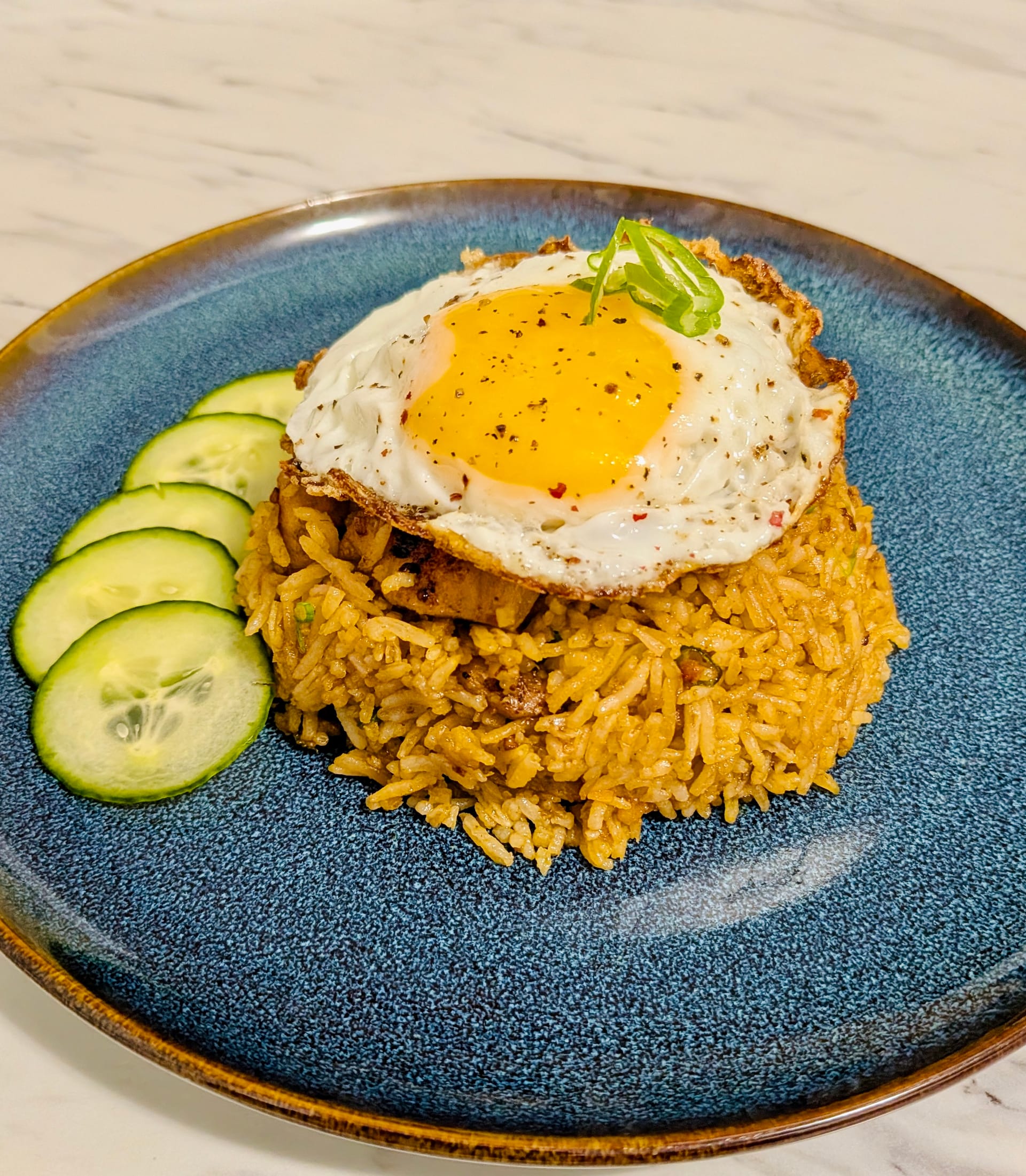 Nasi Goreng fried rice with diced cucumbers on the side and a fried egg on top, on a blue plate.