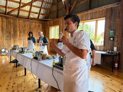 Arnold standing in front of a demonstration table setup with hot plates and pots and sets of ingredients, leading a cooking class as four volunteers stand behind waiting for instructions.