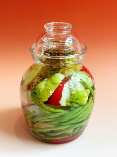 A glass pickling jar filled with long green beans, Sichuan peppercorns, baijiu, chili peppers, and other vegetables on a redish backgrou