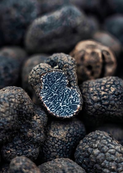 A closeup photo of black truffles. In the center of the pile, one is sliced in half, showing a wavy black and white pattern inside.