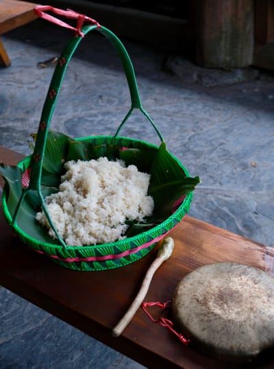 A colorful green and pink basket filled with glutinous white rice sits on a wooden bench.