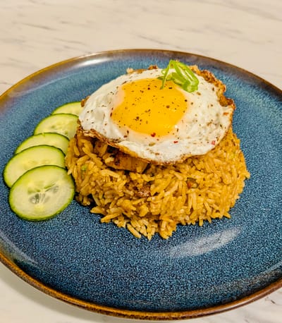 Nasi Goreng fried rice with diced cucumbers on the side and a fried egg on top, on a blue plate.